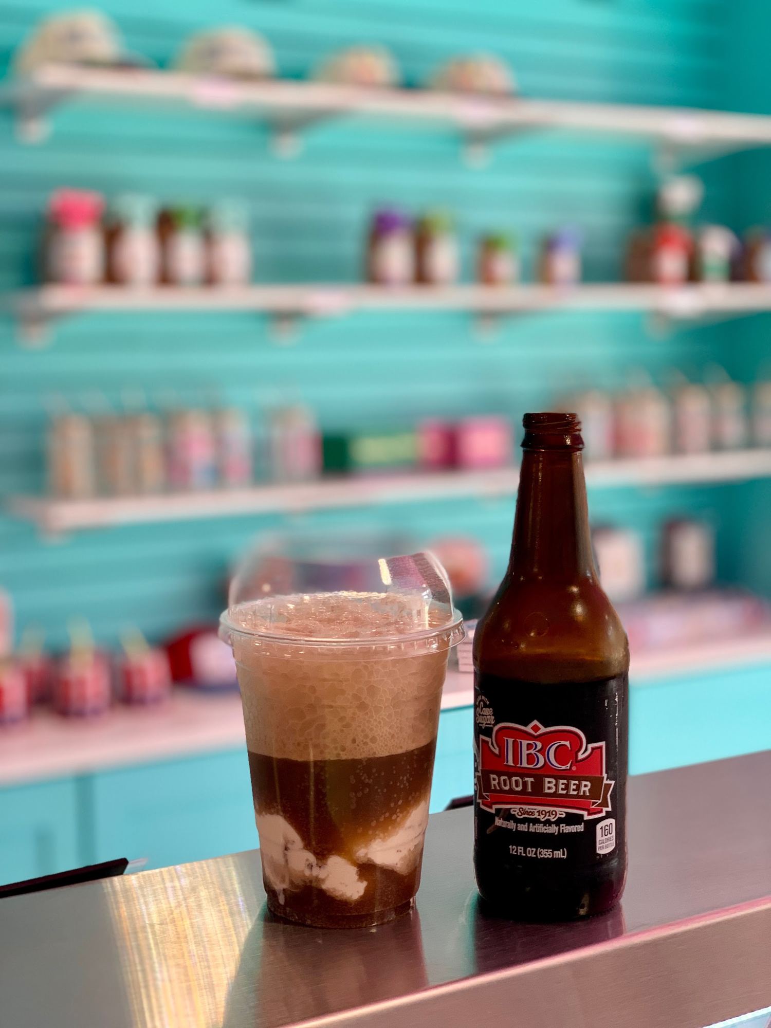 A root beer float in a clear cup next to an IBC root beer bottle, in front of the teal wall of candy jars